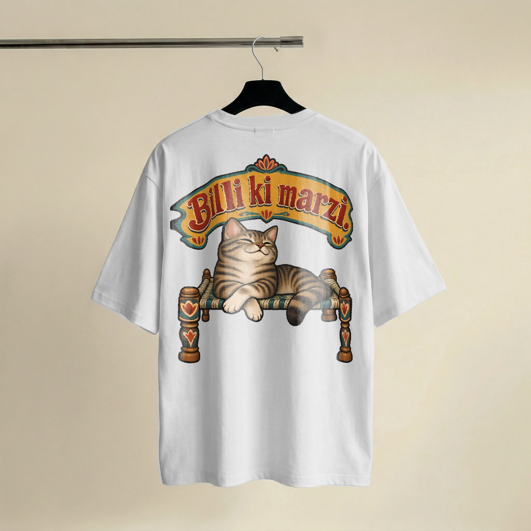 Fat arched signboard lettering reading Billi ki marzi in mustard with red shadowing, above a flat painted tabby cat lounging on a woven charpai with its front paws crossed and eyes half closed, looking very pleased with itself, teal and cream fills with thick black outlines, on a white oversized t-shirt